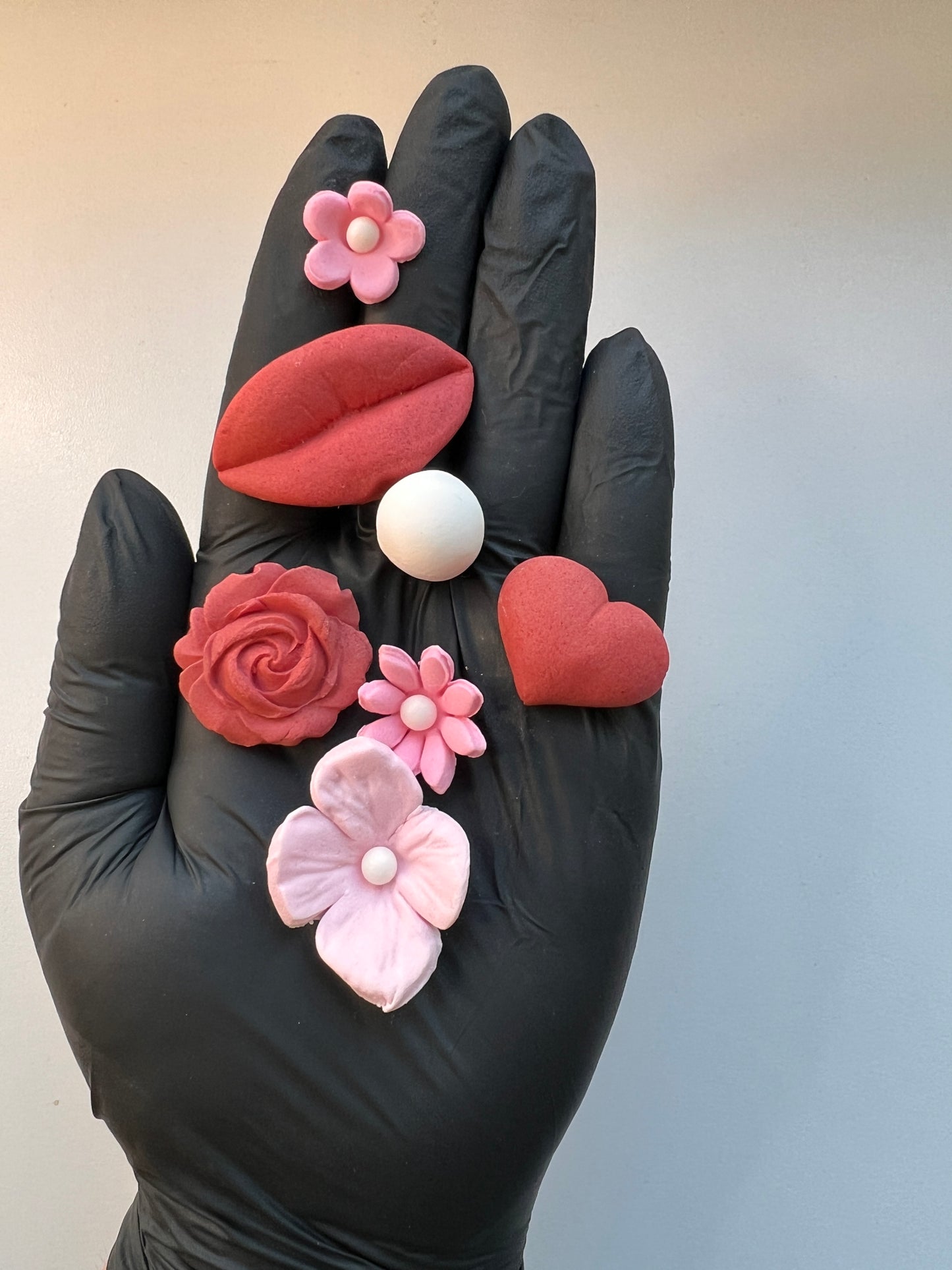 Hand wearing a black glove holding various pink and red fondant Valentine's day decorations against a light background. 