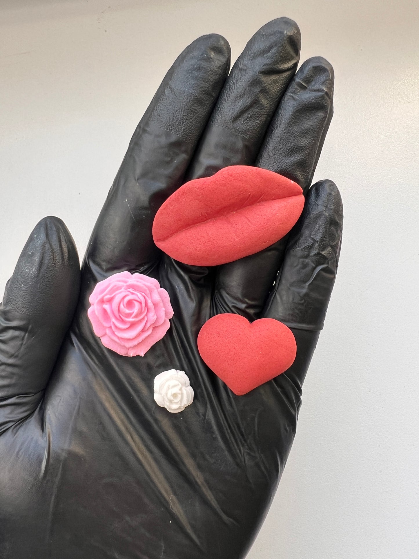 Black-gloved hand holding fondant red heart, pink rose, and white flower against a neutral background