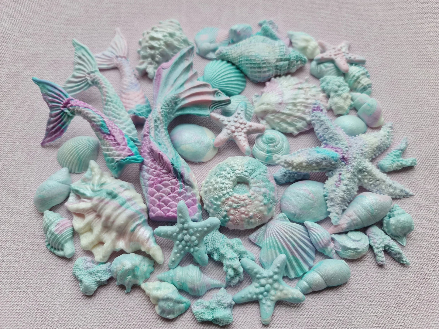 Collection of colorful sea-themed fondant figurines including mermaid tails, starfish, and seashells on a light pink background.