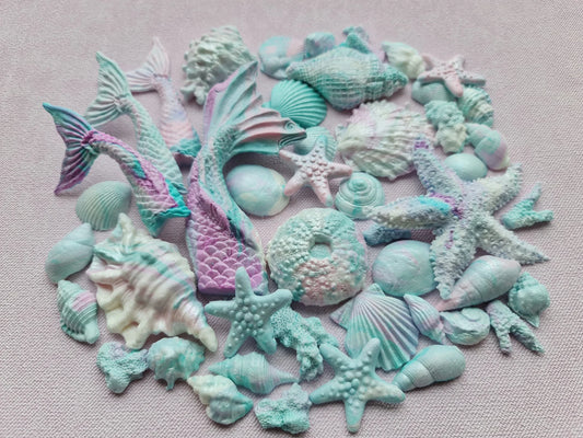 Collection of colorful sea-themed fondant figurines including mermaid tails, starfish, and seashells on a light pink background.