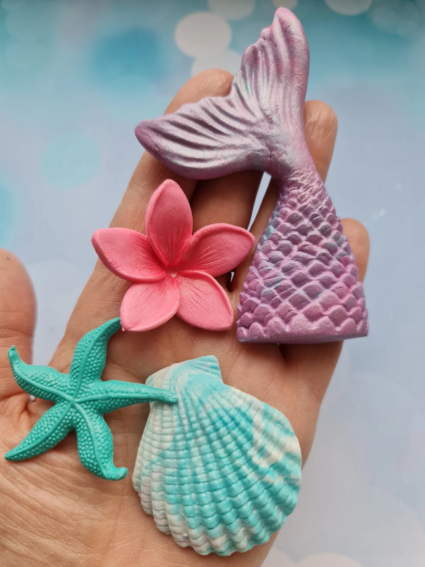 Hand holding fondant items including a pink mermaid tail, pink flower, green starfish, and blue shell against a blurred background.