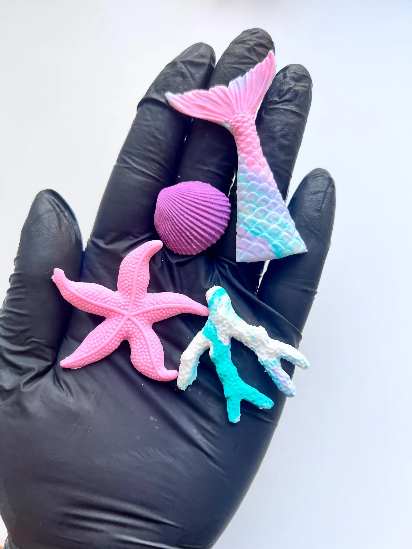 Hand wearing a black glove holding pink and blue mermaid-themed fondant items on a white background